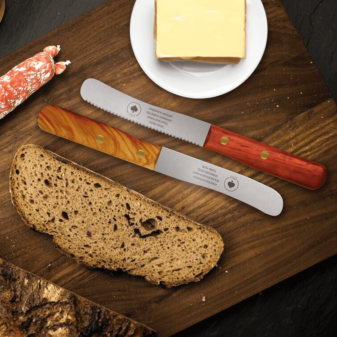 Buckelsmesser und Frühstücksmesser auf einem Holzbrett mit Brot, Käse und Salami für den perfekten Frühstückstisch.
