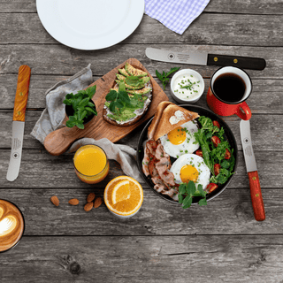 Ein gedeckter Frühstückstisch mit zwei Buckelsmessern, Eiern, Avocado-Toast und Getränken.